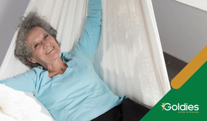 Mulher idosa com cabelos grisalhos cacheados usando um suéter azul claro, relaxando em uma rede branca e sorrindo, ilustrando o post "Os tipos de descanso para melhorar seu bem-estar"