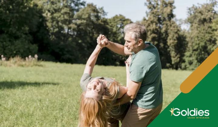 Um homem segura uma mulher brincando enquanto ela se inclina para trás, ambos sorrindo em um campo ensolarado com árvores ao fundo, ilustrando o post "Dança na terceira idade: efeitos no equilíbrio e bem-estar"