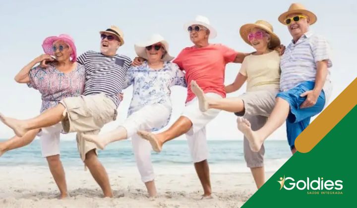 Um grupo de seis adultos mais velhos alegremente chutando as pernas em uma praia, usando chapéus e roupas de verão, ilustrando o post "Os caminhos para a longevidade"