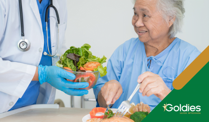 Um profissional de saúde entrega uma salada a uma senhora idosa de vestido azul, que está sorrindo e segurando um garfo, ilustrando o post "A importância dos alimentos para aumentar a imunidade do sênior"