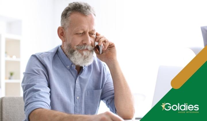 Idoso com barba e cabelos grisalhos fala em um smartphone enquanto olha para um laptop, ilustrando o post "As 5 profissões mais recomendadas para os seniores"